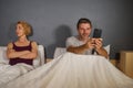 Husband or boyfriend using mobile phone in bed and suspicious frustrated wife or girlfriend feeling upset suspecting betrayal and
