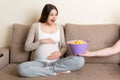 Husband offers his pregnant wife chips relaxing on the sofa at home. Future father takes care about expecting mother