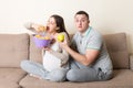 Husband tries to feed his pregnant wife with an apple but she wants chips sitting on the sofa at home. Unhealthy diet during