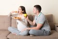 Husband tries to feed his pregnant wife with an apple but she wants croissant sitting on the sofa at home. Unhealthy diet during