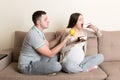 Husband tries to feed his pregnant wife with an apple but she wants a piece of cake sitting on the sofa at home. Unhealthy diet
