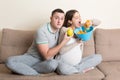 Husband tries to feed his pregnant wife with an apple but she wants potato chips sitting on the sofa at home. Unhealthy diet