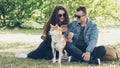 Husband and wife are using smartphone resting on lawn in park, watching screen and talking and fussing dog. Modern