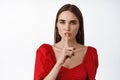 Hush dont speak. Young beautiful woman in red dress tell to keep quiet, taboo gesture, press finger to lips has secret