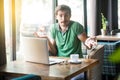 I don`t know! Young confused businessman in green t-shirt sitting with laptop, looking at camera with raised arms and puzzled Royalty Free Stock Photo