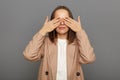 I don`t want to see this. Portrait of young adult Caucasian female wearing beige jacket standing closing her eyes with her palms Royalty Free Stock Photo