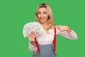 I have big money! Portrait of rich adult woman in stylish overalls pointing at dollar banknotes and smiling