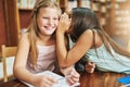 I have a secret. a cheerful young girl whispering into her friends ear while they both sit inside of a classroom during Royalty Free Stock Photo