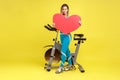 Portrait of woman fitness couch standing near exercise bike and holding big red heart.