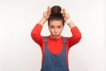 I`m a cow! Portrait of angry girl with hair bun in denim overalls standing with bull horns, antler gesture on her head