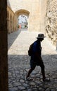 A person silhouette wearing hat and backpack walking down an old street with a stone arch at the background