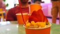 Ice cream with fruit toppings inside orange cup with red spoon, more cups and people out of focus in the background Royalty Free Stock Photo