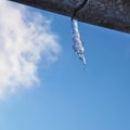 Icicle against blue sky and white cloud. Drop of melted snow falls down. Close up. Icicle hangs from rain gutter on roof. Square Royalty Free Stock Photo