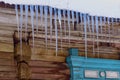 Icicles hang from the roof of a rural wooden house. The ends of the logs and a fragment of the blue trim of the window