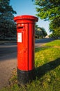 Iconic red british Royal Mail post box in the evening sunshine on a quite road with a green tree lined verge Royalty Free Stock Photo