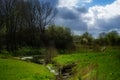Idyllic brook flowing through a green meadow with trees and shrubs, blue sky with clouds in spring, rural natural landscape