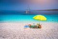 Idyllic white beach with green yellow umbrella on lazy summer day. Sailing boat at anchor in calm crystal clear blue sea