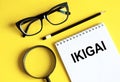 IKIGAI. The word Ikigai on notebook and pencil, magnifier and eyeglasses on yellow background. IKIGAI is a Japanese concept reason