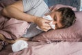 Ill caucasian man in bed with virus, flu or cold blowing running nose surrounded by used tissues