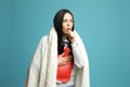 Ill woman with hot water bottle and knitted blanket coughing on light blue background Royalty Free Stock Photo