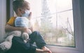 Illness child on home quarantine. Boy and his teddy bear both in protective medical masks sits on windowsill and looks out window