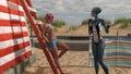 Illustration of a woman in shorts and a halter top leaning against a wood ladder on a beach talking to a blue skin alien in a Royalty Free Stock Photo