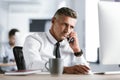 Image of adult businessman 30s wearing white shirt and tie sitting at desk in office by computer, and talking on smartphone Royalty Free Stock Photo