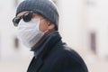 Image of adult man wears shades and protective medical mask, walks outdoor during coronavirus epidemic, looks aside, thinks how to Royalty Free Stock Photo