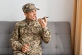 Image of aggressive military man wearing camouflage uniform sitting on sofa, soldier using voice assistant or recording voice