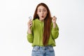 Image of amused redhead girl holds smartphone, looks surprised and curious, receive store notification on mobile phone