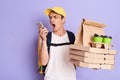 Image of angry emotional young adult man courier in yellow cap and T-shirt holding pizza boxes and coffee to go isolated on purple