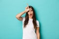 Image of arrogant and sassy brunette woman looking cool, mocking someone, showing loser sign on forehead, standing over