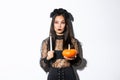 Image of asian woman in wicked witch costume, looking left serious, holding lit candle and pumpkin, celebrating