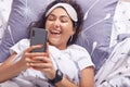 Image of attractive brunette girl lying on pillow with smart phone in hands, wearing t shirt and blindfold, female smiling happily Royalty Free Stock Photo