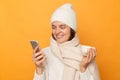 Image of attractive caucasian joyful woman wearing sweater, hat and scarf, holding cup of coffee in hands and using cell phone, Royalty Free Stock Photo