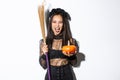 Image of beautiful asian woman dressed-up as a witch for halloween party, holding broom and pumpkin, standing over white Royalty Free Stock Photo