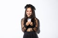 Image of beautiful asian woman in gothic lace dress impersonating witch on halloween party, standing with smartphone and
