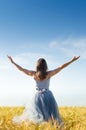 Image of beautiful blond young woman wearing long blue ball dress with arms wide expand looking up on wheat field and blue sky