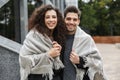Image of beautiful couple man and woman 20s wrapped in blanket, standing over gray building outdoor Royalty Free Stock Photo