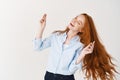 Image of beautiful girl with long red hair snap fingers and dancing, enjoying listening music, standing against white