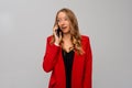 Image of beautiful surprised blonde woman talking on mobile phone, calling on cellphone, standing in red blazer against gray Royalty Free Stock Photo