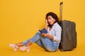 Image of beautiful woman with gray suitcase, holding passport and tickets in hand, posing isolated over yellow background, sitting Royalty Free Stock Photo