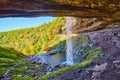 Behind huge waterfall tucked into cliffs with open view of fall forest mountains and small tourists below Royalty Free Stock Photo