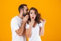 Image of brunette man whispering secret or interesting gossip to woman in her ear, isolated over yellow background
