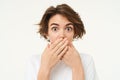 Image of brunette woman shuts her mouth, looks surprised, puts hands over her lips, stands over white background