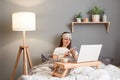 Image of calm smiling woman wearing sleeping mask sitting in bed with laptop, holding cell phone, checking social networks or