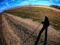The image captures the shadow of an individual holding a stick, standing before an open field with a lone tree and distant woods