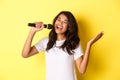 Image of carefree african-american girl singing in microphone, smiling happy, performing over yellow background Royalty Free Stock Photo