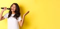 Image of carefree african-american girl singing in microphone, smiling happy, performing over yellow background Royalty Free Stock Photo