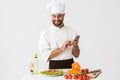 Image of caucasian chef man in uniform smiling and holding smartphone while cooking vegetable salad